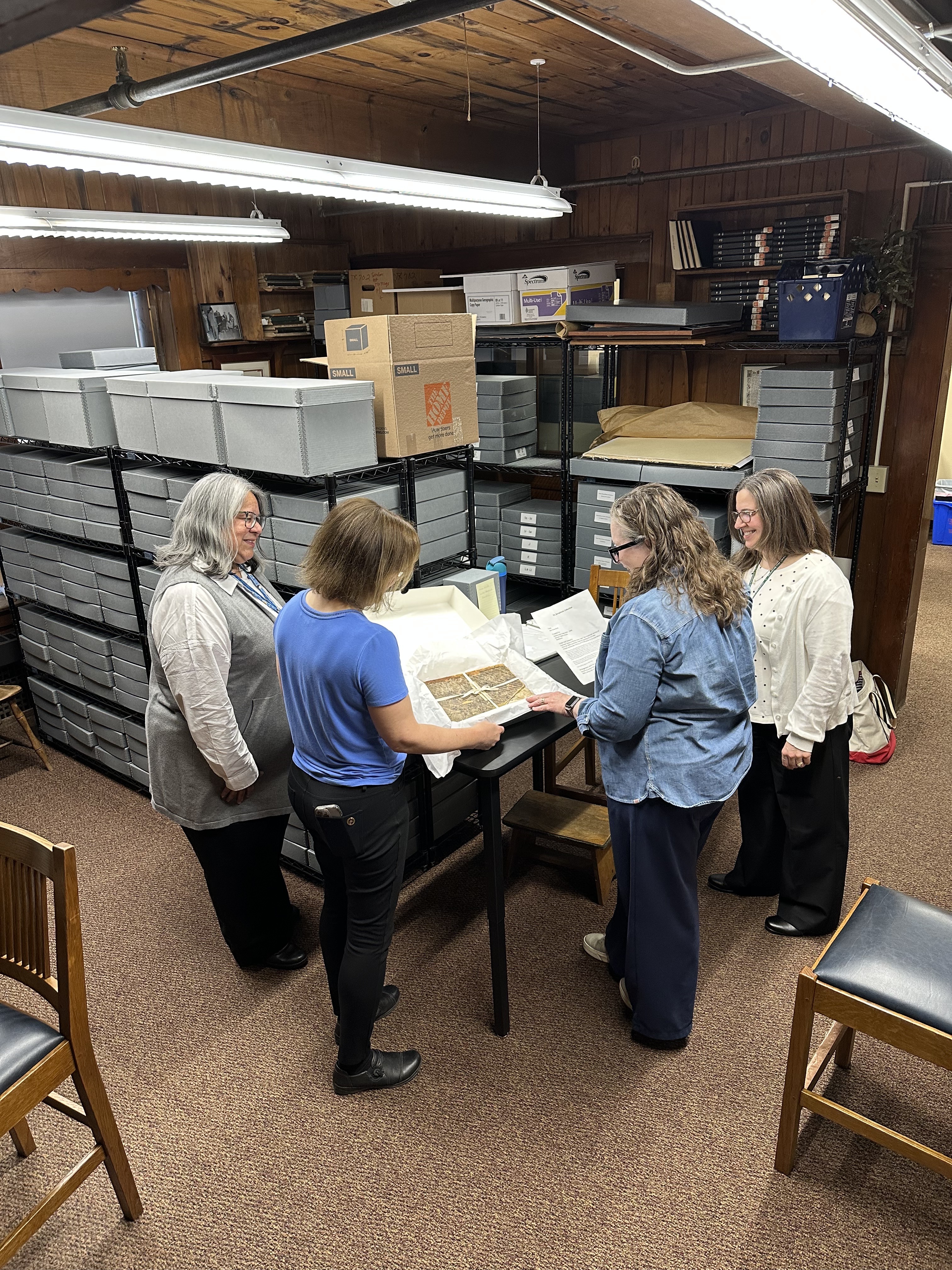 Four people stand around table in an archive. The two in the middle are holding open a box, which holds an old book wrapped in tissue paper.