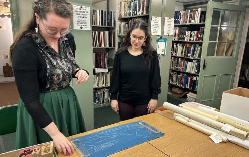 Two individuals stand at a wooden table reviewing a blueprint.