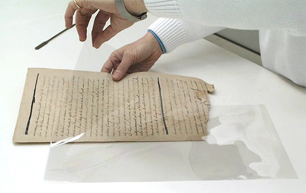 Two bare hands placing a fragile, handwritten vintage document into a clear polyester enclosure.  